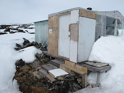 L’entrée d’une cabane où la neige semble avoir fondu; elle est épaisse tout autour. Un escalier de morceaux de planches est déposé sur des pierres entassées. La cabane est un assemblage de morceaux de bois et de tôle; certains sont couverts de peinture pâle délavée.