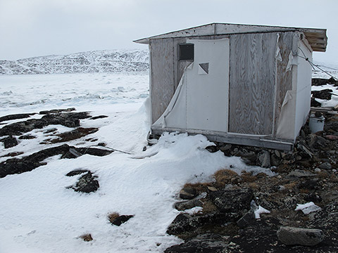 Sur les rivages rocheux et partiellement couverts de neige d’une crique gelée, une cabane repose sur un amoncellement de pierres. Elle est composée de panneaux de bois usés. Certains bouts d’un ancien recouvrement de toile pendent, déchirés.