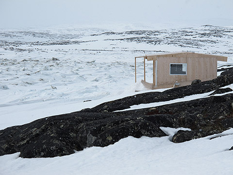 Dans un paysage vallonné et rocheux, principalement couvert de neige et de glace, une cabane de bois semble récemment installée. Elle est composée de panneaux de bois de teinte naturelle pas encore grisonnés par le soleil.