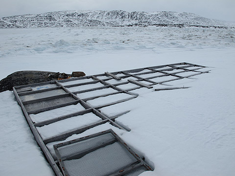 Sur les rivages d’une crique gelée bordée d’une montagne rocheuse partiellement couverte de neige, une plateforme de bois commence à apparaître en raison de la fonte de la neige qui la recouvrait. Cette plateforme est divisée en caissons rectangulaires. Un cadre grillagé repose sur la plateforme à l’avant-plan.