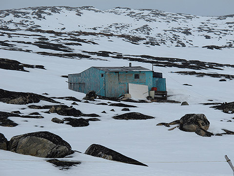 Dans une colline rocheuse partiellement couverte de neige se trouve une cabane. Elle est composée de panneaux de bois usés dont la peinture bleutée est délavée. Quelques objets et matériaux de construction sont déposés autour de la cabane.