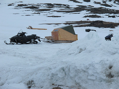 Un traîneau couvert est attaché à une motoneige. Il est fabriqué de morceaux de panneaux de bois et de tôles. Il représente un petit habitacle fermé sur skis. Tout autour, il y a de la neige au sol. Une autre motoneige et un toboggan reposent tout près. Deux véhicules tout-terrain sont révélés par la neige qui fond. Ils sont encore semi-enterrés. En arrière-plan, on aperçoit une colline rocheuse partiellement couverte de neige.